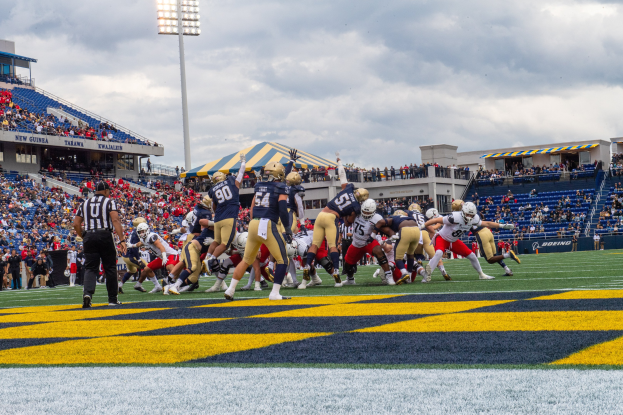 Fußballspieler in Sportkleidung und Helmen konkurrieren auf einem Feld, mit Zuschauern und Stadionstrukturen im Hintergrund.