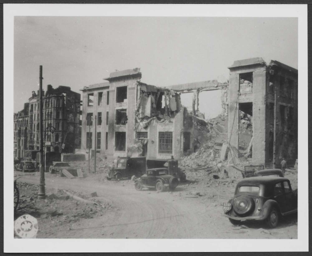 Ein Schwarz-Weiß-Foto einer Straße mit parkenden Autos vor einem zerbombten Gebäude, wahrscheinlich nach einem Bombenangriff in Berlin, Deutschland, in einem Rahmenformat angezeigt.