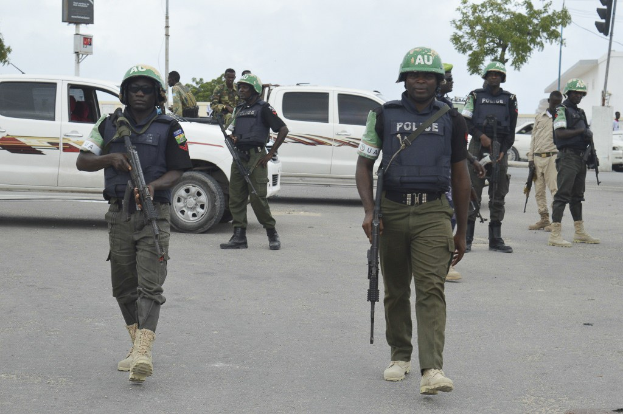 Eine Gruppe von Polizisten in Helmen und taktischer Ausrüstung geht eine Straße entlang, bewaffnet mit Fahrzeugen, Verkehrszeichen, Bäumen, Gebäuden und einem bewölkten Himmel im Hintergrund.