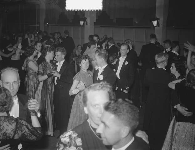 Eine Gruppe von Menschen, die auf einer Party in einem Schwarz-Wei├č-Setting tanzen, wobei einige Gl├Ąser halten und eine beleuchtete Wand im Hintergrund zu sehen ist.