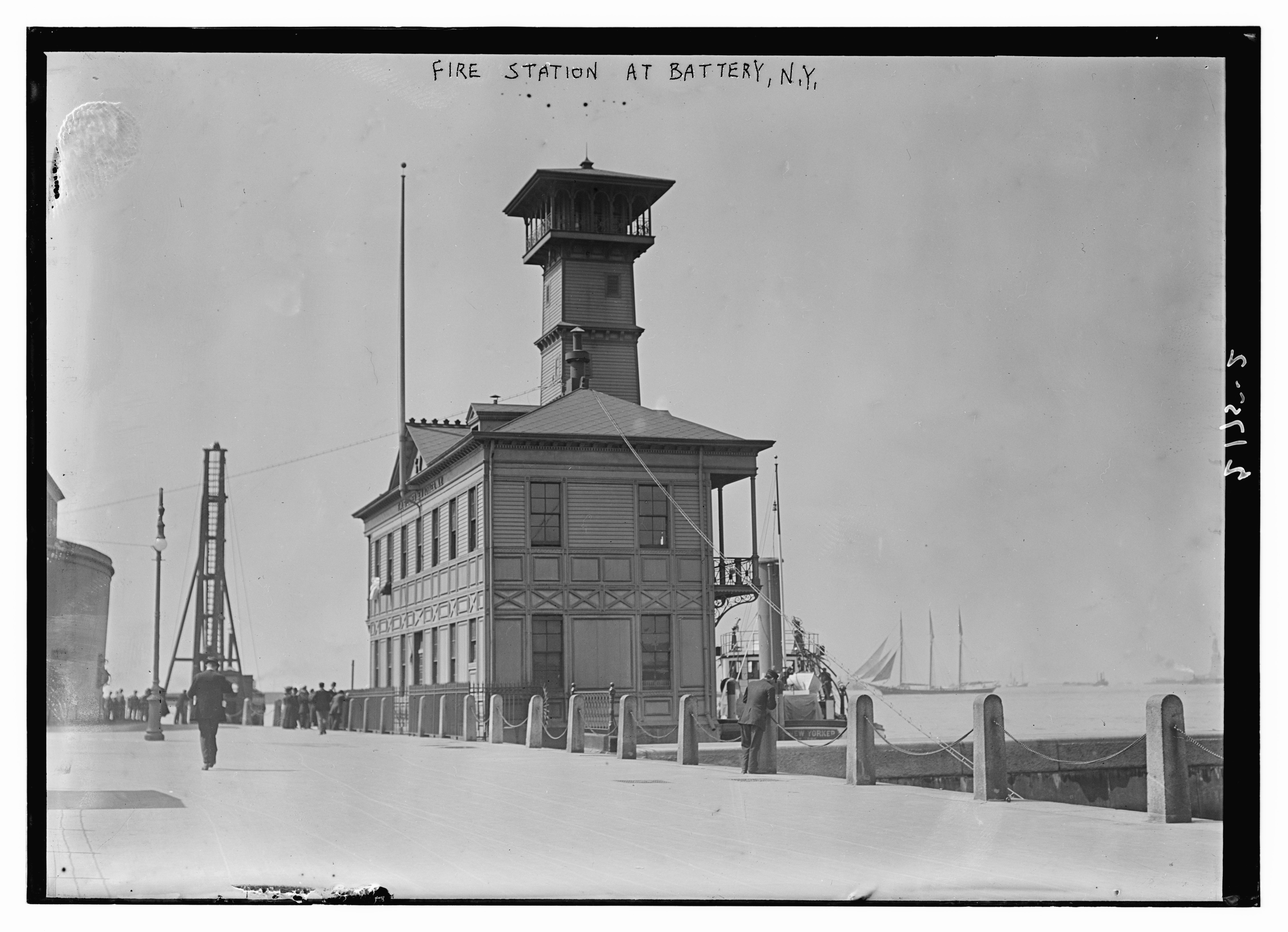 Ein Schwarz-Weiß-Foto einer Feuerwache in Battery, New York, mit einem Gebäude, Menschen, Pfählen, einem Zaun, Booten auf dem Wasser und einem bewölkten Himmel.