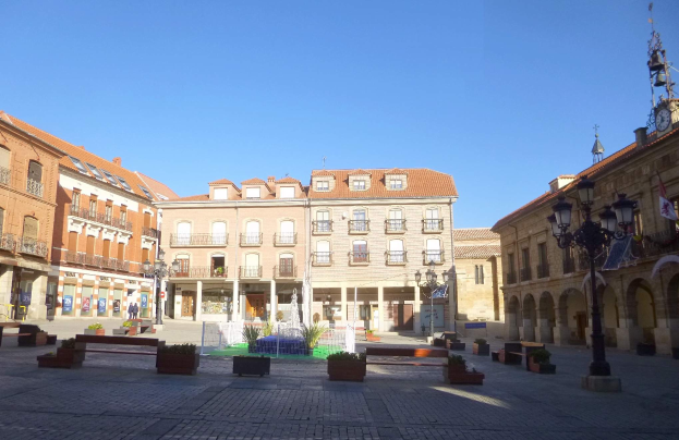 Ein Plaza Mayor in einem Stadtplatz mit einem zentralen Brunnen, umgeben von Bänken, Topfpflanzen, Straßenmasten, Straßenlaternen, einem Uhrenturm und Gebäuden mit Fenstern unter einem klaren blauen Himmel.