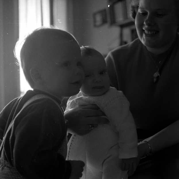 Eine Frau lächelt warm, während sie ein Baby hält, mit zwei anderen Kindern neben ihr; gerahmte Bilder und ein Fenster sind im Hintergrund zu sehen.
