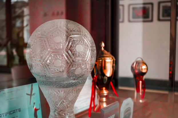 Eine Glastisch zeigt eine zentrale Glastrophäe zwischen anderen Trophäen und einem Zertifikat, mit gerahmten Gegenständen an der Wand im Hintergrund.