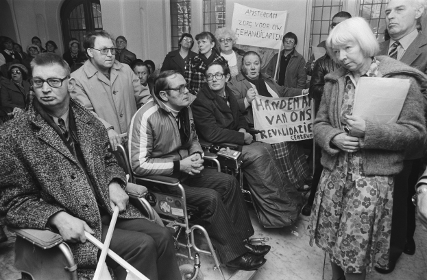 Eine Gruppe von Menschen in Rollstühlen und Stehenden, die Schilder halten, vor einer Menge, mit Fenstern und einer Wand im Hintergrund, in Schwarz-Weiß.