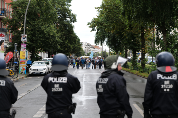 Eine Gruppe von Polizisten in schwarzen Uniformen und Helmen steht an der Straße, mit Menschen, Fahrzeugen, Fahrrädern, Pfählen, Plakaten, Bäumen, Gebäuden und einer Banner mit Text im Hintergrund.