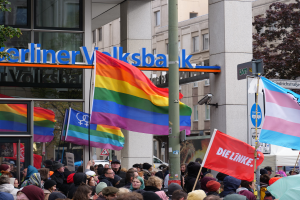 Eine große Gruppe von Menschen mit Fahnen und Transparenten steht vor einem Gebäude, mit einem Mast im Vordergrund und Bäumen auf beiden Seiten, bei einer Gay-Pride-Parade in Berlin.
