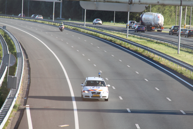 Ein Auto fährt auf einer Autobahn mit einem Polizeiwagen am Straßenrand, anderen Fahrzeugen, Leitplanken, Pfählen, Planken und Bäumen im Hintergrund.