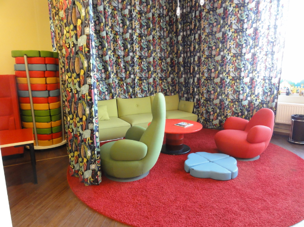 Ein Wohnzimmer mit Sofas, Stühlen und einem Tisch auf einem roten Teppich, mit einem Buch auf dem Tisch, einem Kissen auf dem Sofa, einem bunten Vorhang und verschiedenen Gegenständen an der Wand im Hintergrund.