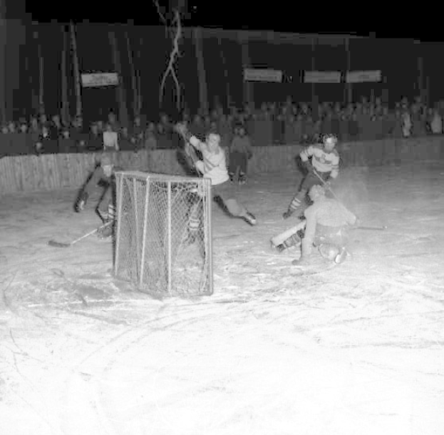 Schwarzes und weißes Foto eines Hockey-Spiels im Gange auf einem Eisstadion, mit Spielern in der Nähe eines Tors und Zuschauern im Hintergrund.