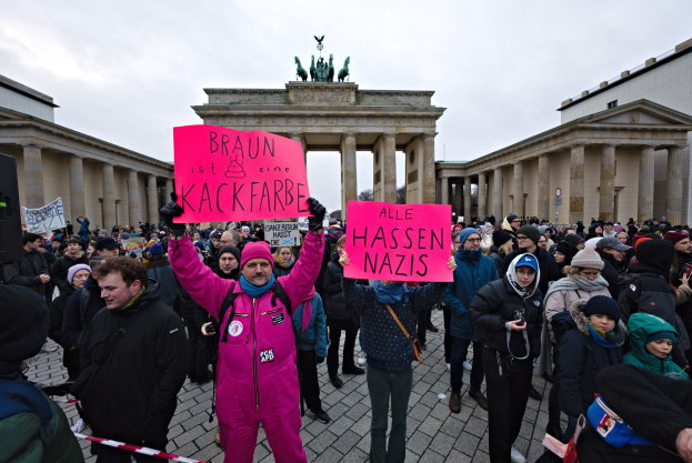 Eine Gruppe von Menschen protestiert vor dem Reichstagsgebäude in Berlin, hält Schilder gegen die Regierungskampagne gegen Neonazis hoch, mit Gebäuden, Säulen und Statuen im Hintergrund unter einem klaren blauen Himmel.