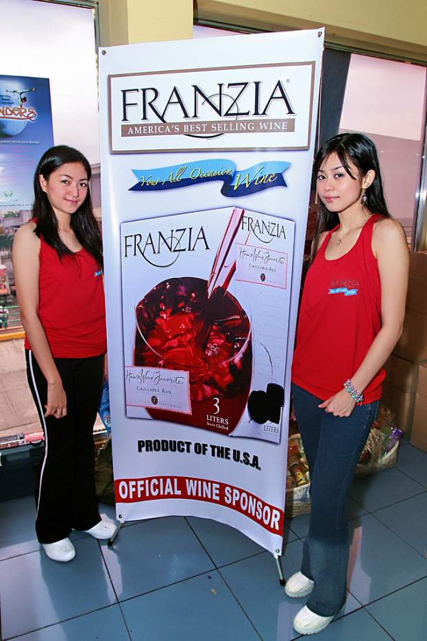 Zwei Frauen neben einem Plakat für Franzia, Amerikas bestverkauften Wein, mit einem Glasfenster im Hintergrund.