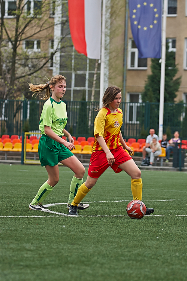 Zwei Frauen spielen Fußball auf einem Feld, mit einem Ball zwischen ihnen, während vier Personen auf Stühlen im Hintergrund sitzen, mit einem Gebäude und Fahnen dahinter.