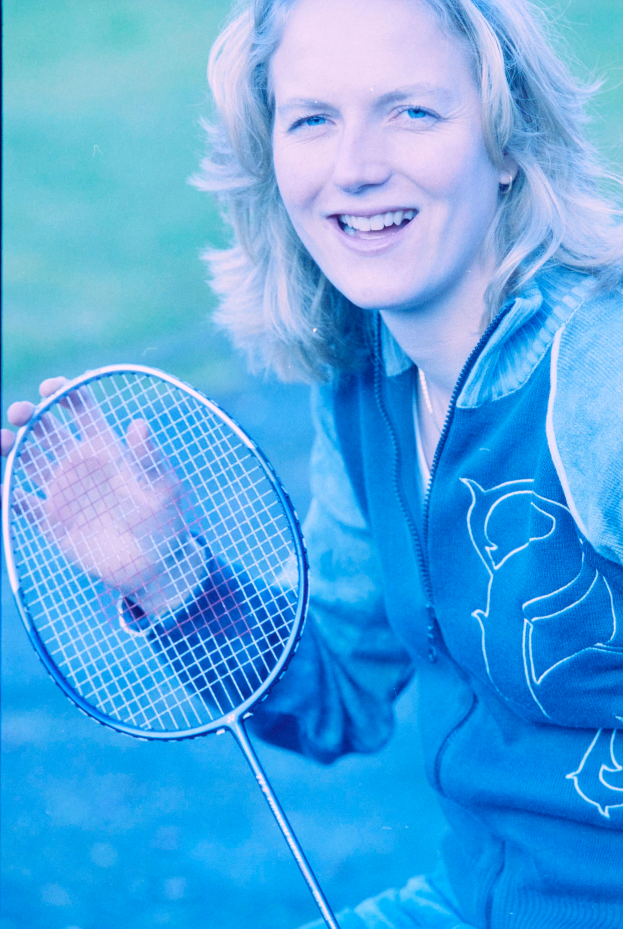 Eine Frau mit strahlendem Lachen, die ein weißes Hemd und blaue Shorts trägt, hält einen Badmintonschläger in der Hand und schaut konzentriert darauf.