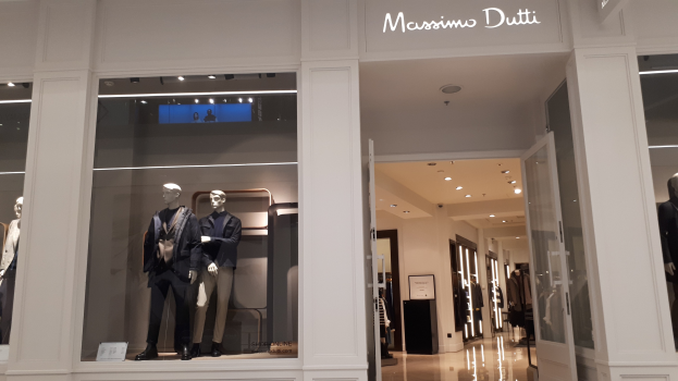 Vitrine des Massimo Dutti in Mailand, Italien, mit Schaufensterpuppen in moderner Kleidung, beleuchteter Decke, Informationsschildern und sichtbarem Boden.