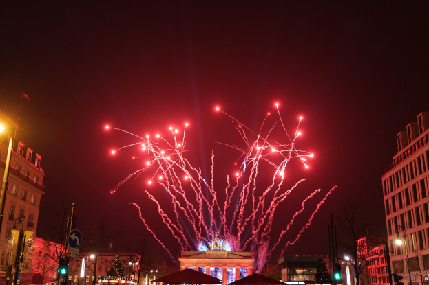 Eine belebte Stadtstraße bei Nacht am Silvesterabend in Berlin, mit Gebäuden, Bäumen, Laternen, Ampeln, Schildern, Zelten, Menschen und einem beeindruckenden Feuerwerk am Himmel.