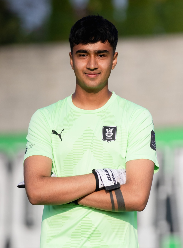 Ein junger Mann in einem grünen T-Shirt steht auf einem Fuballfeld und hat die Arme verschränkt, lächelt, wahrscheinlich ein Torwart, mit einem unscharfen Hintergrund.