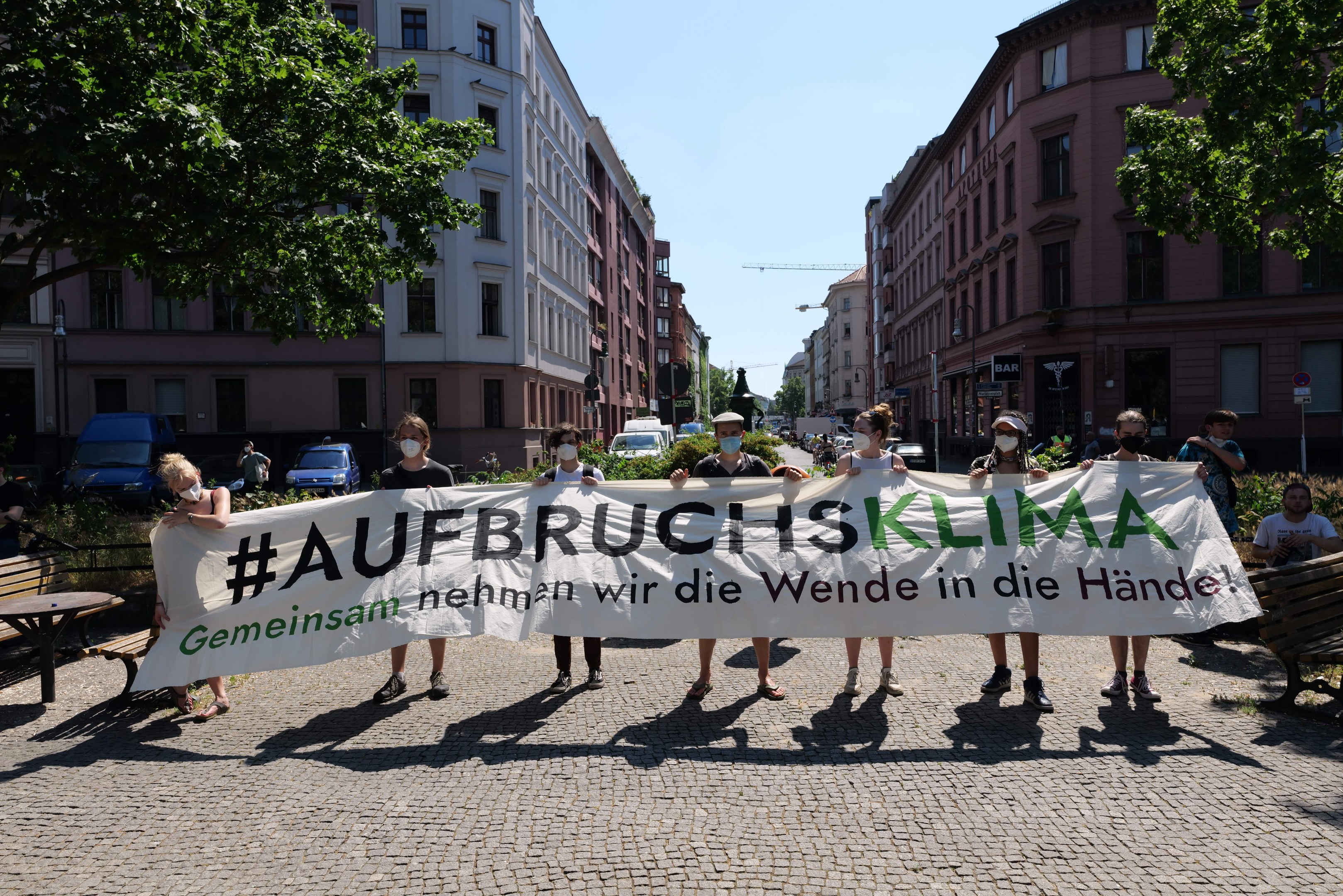 Eine Gruppe von Menschen mit Masken, die ein Banner mit der Aufschrift 'Aufbruchsklima' tragen, während sie auf einer Klimademonstration in Berlin, Deutschland, vor einem Gebäude mit Außenbestuhlung und Grünflächen protestieren.