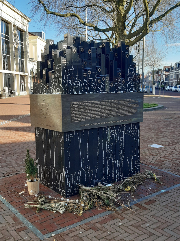 Mahnmal für Holocaust-Opfer in einem Stadtplatz mit einem Baum davor, einem Topfgewächs, verstreuten trockenen Blättern und Gebäuden im Hintergrund.