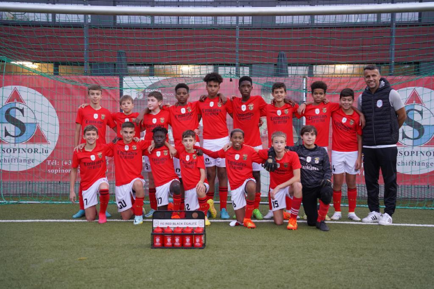 Eine Gruppe junger Männer in roten T-Shirts und schwarzen Shorts steht auf einem Fußballfeld vor einer Kiste roter Gegenstände, im Hintergrund Schilder mit der Aufschrift "FC Rot-Schwarz Eglaute - Champions League".