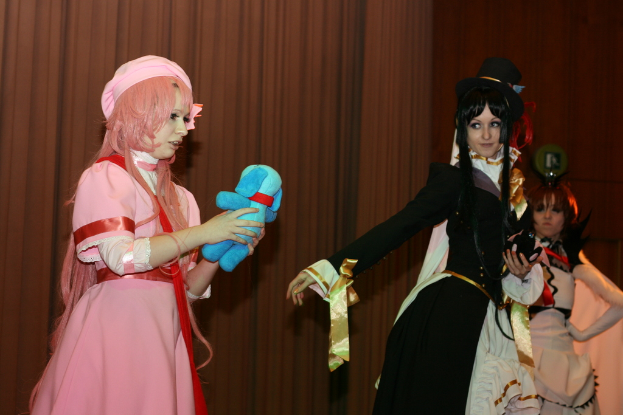 Drei Frauen sind in einer dramatischen Performance zu sehen, wobei eine in einem pinken Kleid einen blauen Teddybär hält, eine andere in einem schwarzen Kleid nach links schaut und ein brauner Vorhang im Hintergrund zu sehen ist.