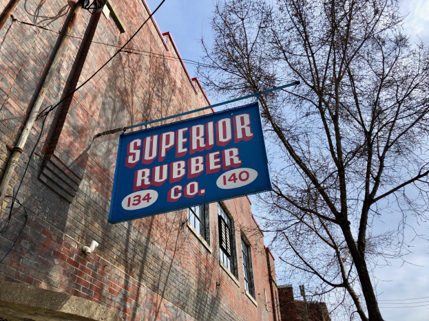 Ein Gebäude mit Fenstern, Rohren und einem Schild mit der Aufschrift "Superior Rubber Co" ist zu sehen, rechts daneben ein Baum und im Hintergrund ein klarer blauer Himmel.