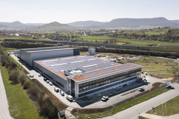 Luftaufnahme eines große Gebäudes mit Solarpanelen auf dem Dach, umgeben von Fahrzeugen, Bäumen, Gras und Masten, mit Höhen und einem klaren blauen Himmel im Hintergrund.