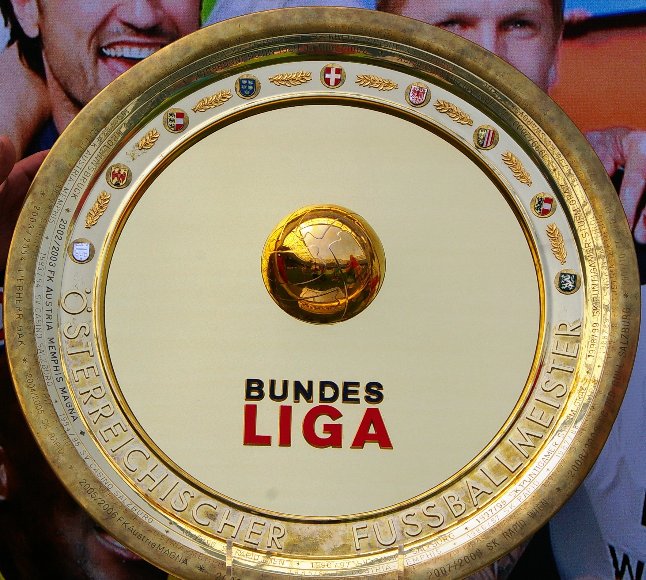 Goldener Schild mit "Bundesliga"-Text, gehalten von einem Mann mit einem Banner im Hintergrund, das drei Personen zeigt.