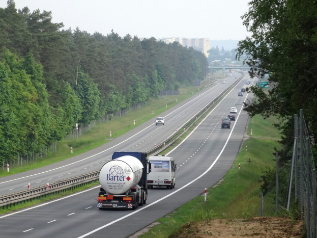 Ein großer Lkw fährt auf einer von beiden Seiten von Bäumen gesäumten Autobahn, mit einem Zaun und Gras auf der rechten Seite und im Hintergrund sichtbare Gebäude unter einem klaren blauen Himmel.