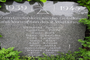 Ein Gedenkstein mit einem Kreuz, umgeben von gr├╝nen Pflanzen, mit Inschrift, wahrscheinlich zur Erinnerung an Kriegsopfer.