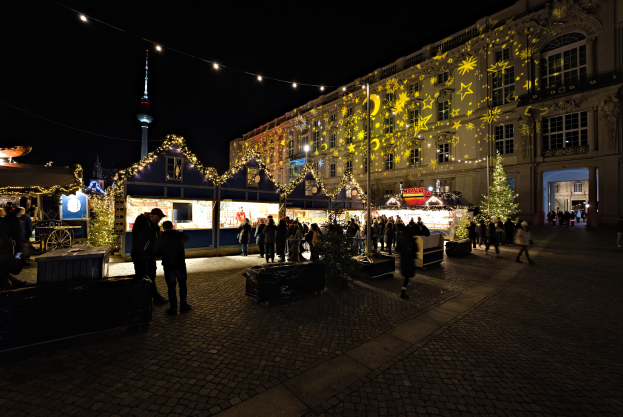 Ein lebendiger Weihnachtsmarkt in Berlin, Deutschland, mit Menschen um beleuchtete und dekorierte Stände, Gebäuden mit Fenstern im Hintergrund unter einem dunklen Himmel.