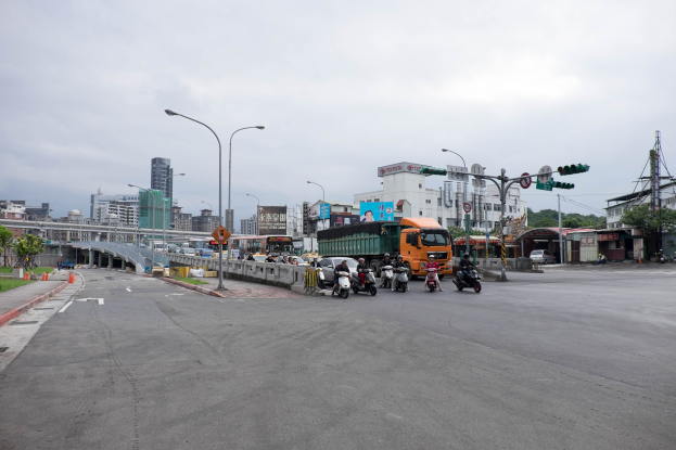 Eine belebte Stadtstraße mit verschiedenen Fahrzeugen, darunter ein Lastwagen und ein Auto, Laternenpfähle, Verkehrsampeln, Schilder, eine Brücke mit Geländern, Bäume, Gebäude mit Fenstern und ein bewölkter Himmel.