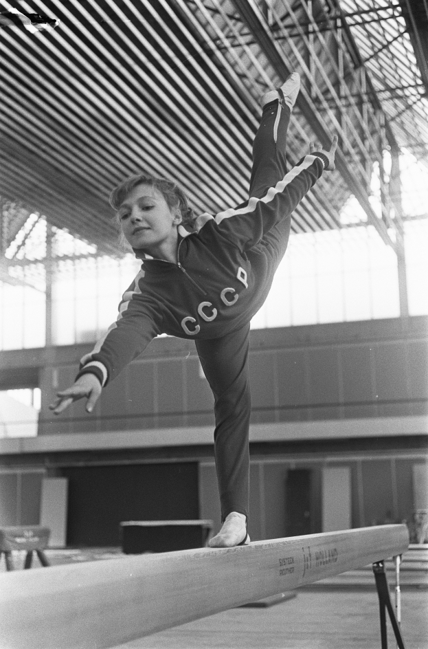 Ein junges Mädchen zeigt Gymnastik auf einem Balancebalken in einer Turnhalle, mit einer Wand und einer Decke mit Stangen im Hintergrund.