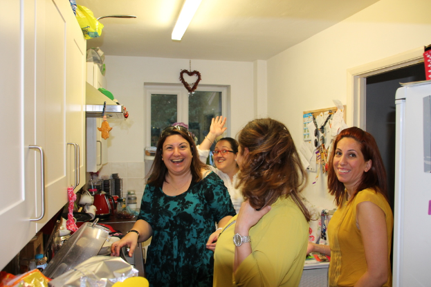 Eine Gruppe strahlender Frauen in einer Küche mit Kühlschrank, Gefäßen auf der Arbeitsplatte, Schränken und einer Wandtafel, wobei eine Frau eine Brille trägt und eine Lampe an der Decke.