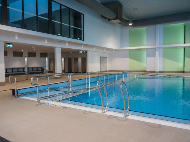 Indoor-Schwimmbad mit gefliestem Boden, Metallstangen, Säulen, Türen, Lampen und Decke in einem großen Gebäude.