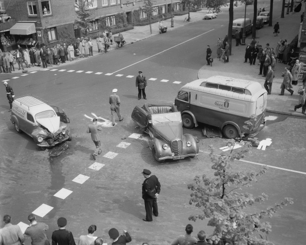 Schwarz-weiß-Szene eines Autounfalls an einer Straßenkreuzung mit mehreren Fahrzeugen, darunter ein Lieferwagen, und einer Gruppe von Zuschauern; Gebäude, Bäume, Strommasten und Texttafeln im Hintergrund.