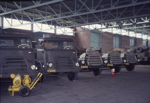 Eine Gruppe von Lastwagen, die in einem Gebäude mit einem sichtbaren Feuerlöscher, Metallstangen, Fenstern, Deckenlampen und Rädern an den Lastwagen geparkt sind.