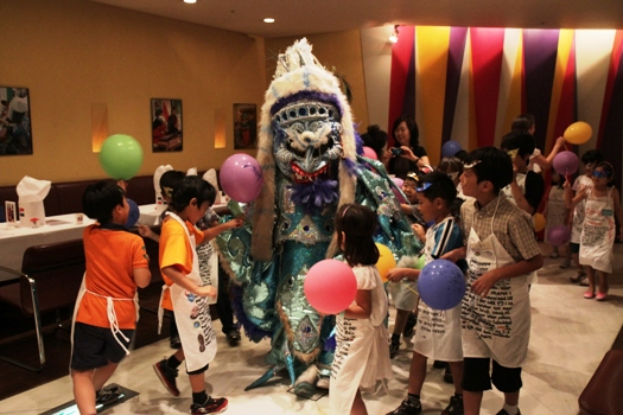 Eine Gruppe von Kindern umringt eine Person in einem Kostüm, die Ballons hölt, mit einer Frau, die Fotos macht, im Hintergrund, in einem Raum mit Tischen, Stühlen, Rahmen und Deckenleuchten.