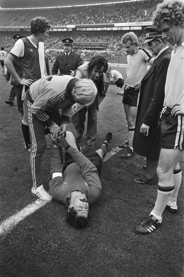 Schwarz-weißes Bild eines Fußballspielers auf dem Boden liegend, umgeben von einer Gruppe von Männern, mit einer Menge im Hintergrund eines Stadions.