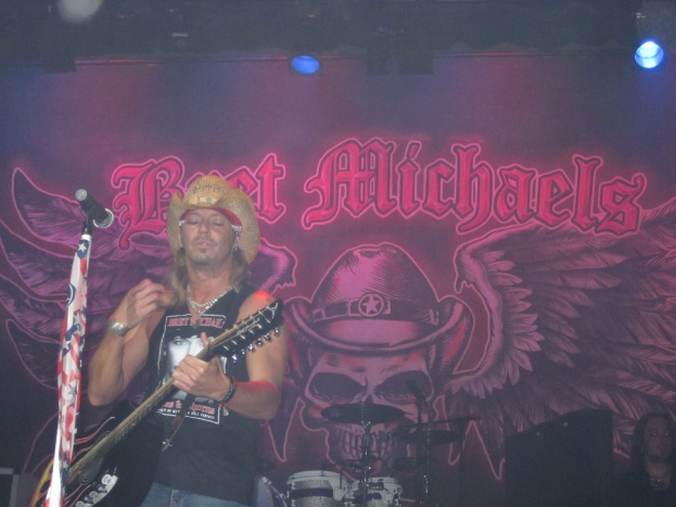 Eine Musik-Konzertszene mit einem Gitarristen auf der Bühne, einer Person in der unteren rechten Ecke, einem Banner im Hintergrund und Drogen in der Mitte unten, mit Scheinwerfern oben.