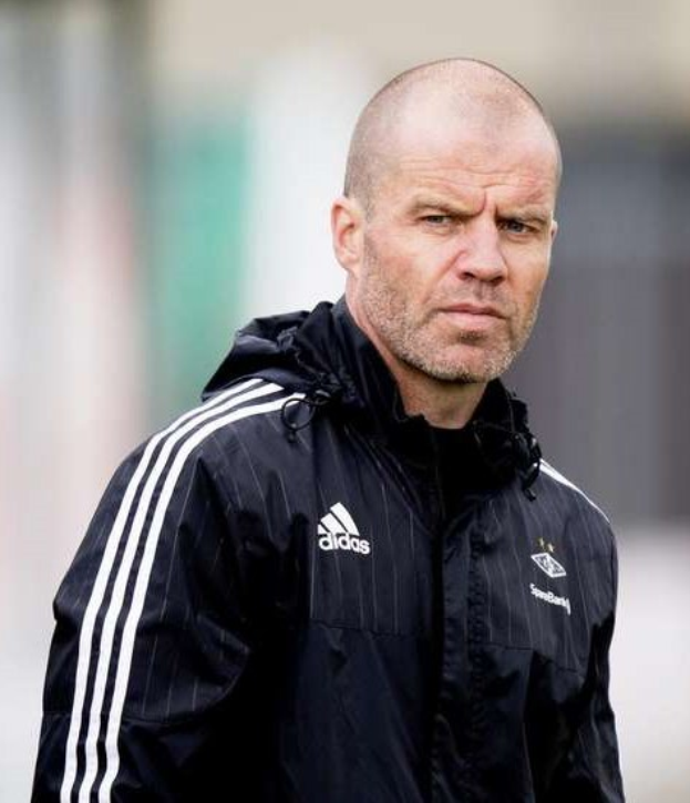 Bald Trainer in marineblauer Jacke mit weißen Streifen und weißem Kragen, der auf einem Fußballfeld steht und die Hände verschränkt hält.