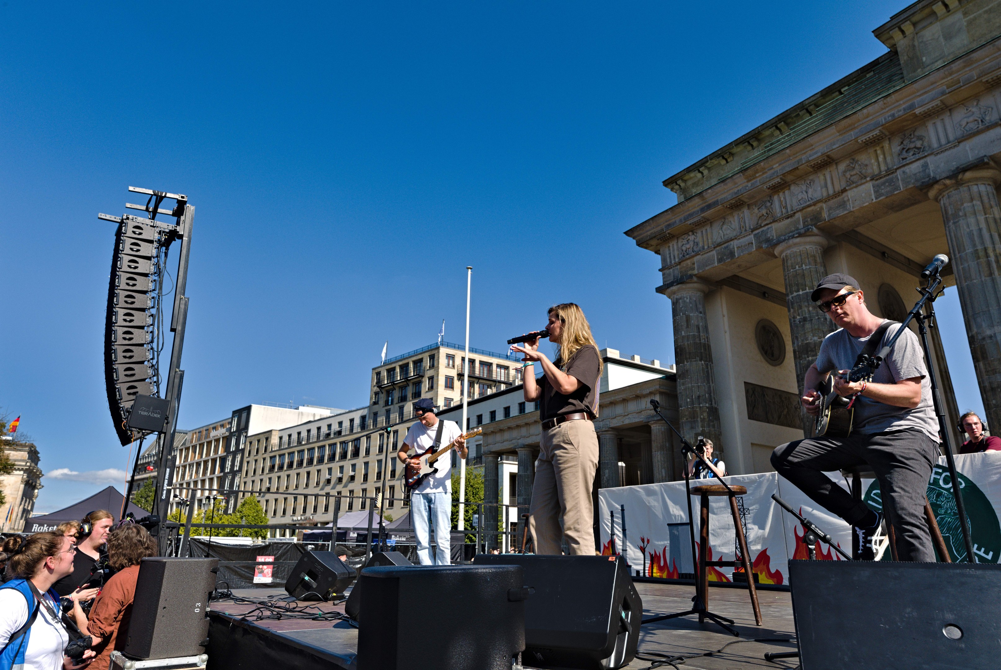 Eine Gruppe von Menschen, die auf einer Bühne vor dem Brandenburger Tor in Berlin Musik spielen, umgeben von Lautsprechern und Equipment, vor einem klaren blauen Himmel.