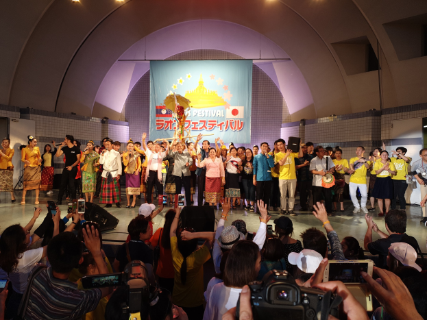 Gruppe von Menschen auf einer Bühne mit Kameras, Handys und Musikinstrumenten während einer Festivaleröffnung mit Banner, Wänden und Deckenlampen im Hintergrund.