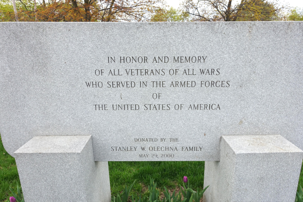 Gedenkstein mit Inschrift für US-Streitkräfte-Veteranen, umgeben von Pflanzen, Blumen, Gras, Bäumen und einem bewölkten Himmel.