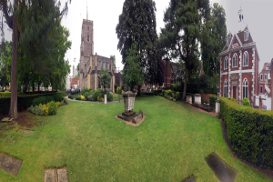 Ein Park mit einem Brunnen in der Mitte, umgeben von Gras, Pflanzen, Bäumen und Gebäuden mit Fenstern, Fahrzeugen auf der Straße und einem klaren blauen Himmel, mit einem prominenten Baum in der Mitte.