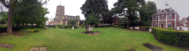 Ein Park mit einem Brunnen in der Mitte, umgeben von Gras, Pflanzen, Bäumen und Gebäuden mit Fenstern, Fahrzeugen auf der Straße und einem klaren blauen Himmel, mit einem prominenten Baum in der Mitte.