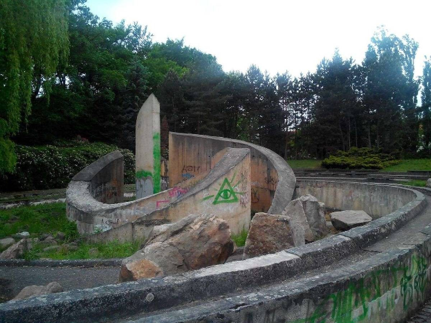 Verlassener Skatepark mit graffiti-bedeckten Wänden, umgeben von Felsen, Gras, Pflanzen und Bäumen unter einem klaren blauen Himmel.