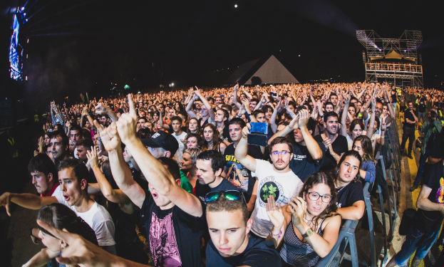 Eine große Menge Menschen auf einem Musikfestival mit erhobenen Händen, einige sitzen und andere stehen, begrenzt von einem Metallzaun auf der rechten Seite, mit Gebäuden und Lichtern im Hintergrund unter einem dunklen Himmel.