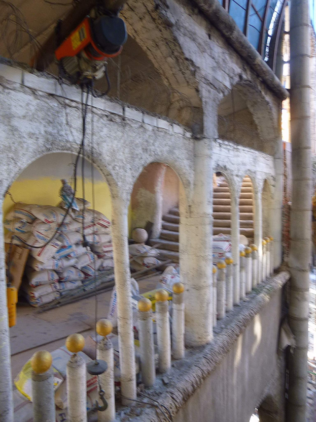 Altes Gebäude mit Bogengang und Säulen wird renoviert, mit verstreuten Werkzeugen, Materialien und Gegenständen wie Taschen, Geländern und einer Maschine mit Drähten oben drauf.