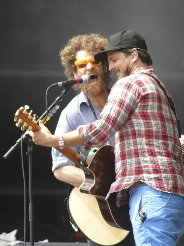 Zwei Männer mit Gitarren auf der Bühne, einer in einem rot-blau karierten Hemd mit blauen Hosen, der andere mit einer Sonnenbrille und einer schwarzen Mütze.